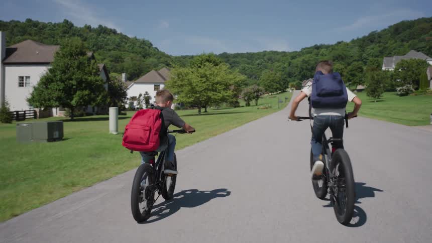 Two boys ride bicycles down a sunny suburban street toward the hills