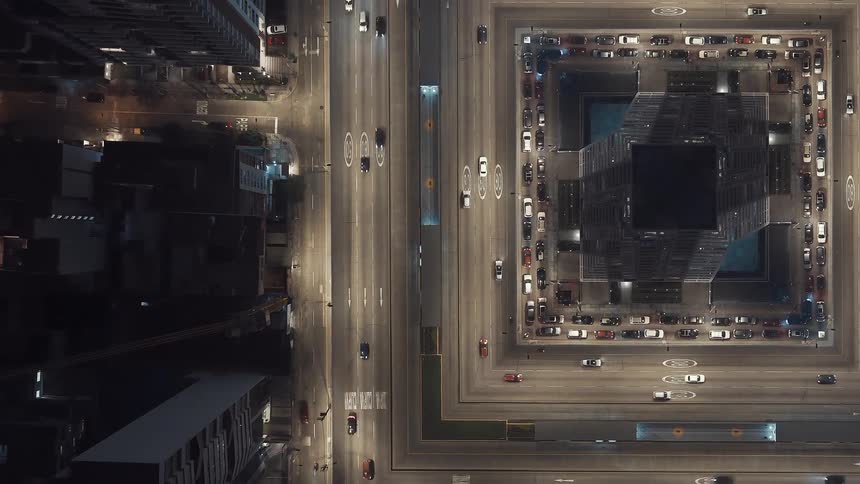 This is a highangle birdseye view of a city street and a large symmetrical building complex at night the main street running vertically has busy traffic moving in both directions marked by bright white headlights and red taillightson the right