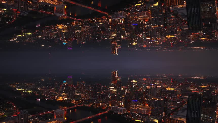 A vertically mirrored aerial view pulls back over an illuminated city skyline at night