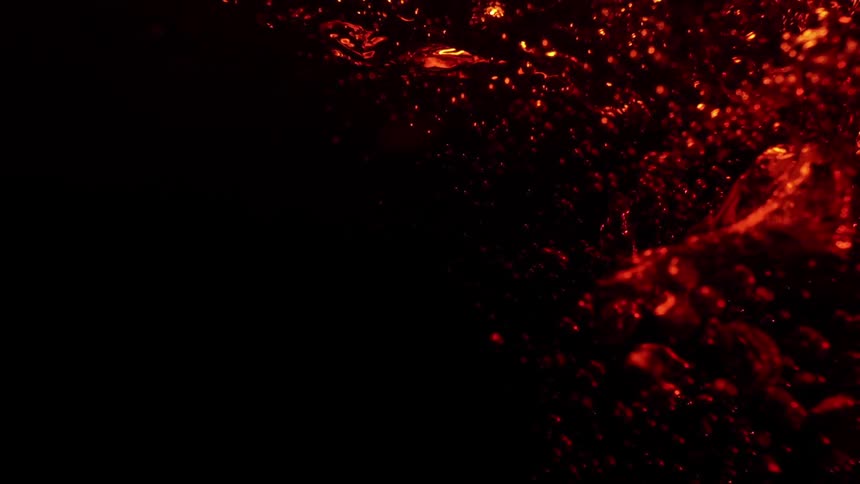 A mass of intensely illuminated red or orange liquid moves and splashes against a completely black background creating numerous bright droplets and streaks that scatter into the dark space