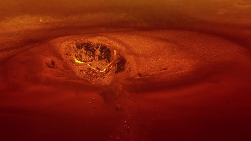An extreme closeup shot focuses beneath the surface of a carbonated orangeamber liquid where a stream of liquid is pouring in creating a dynamic pocket of turbulent foam and rising bubbles