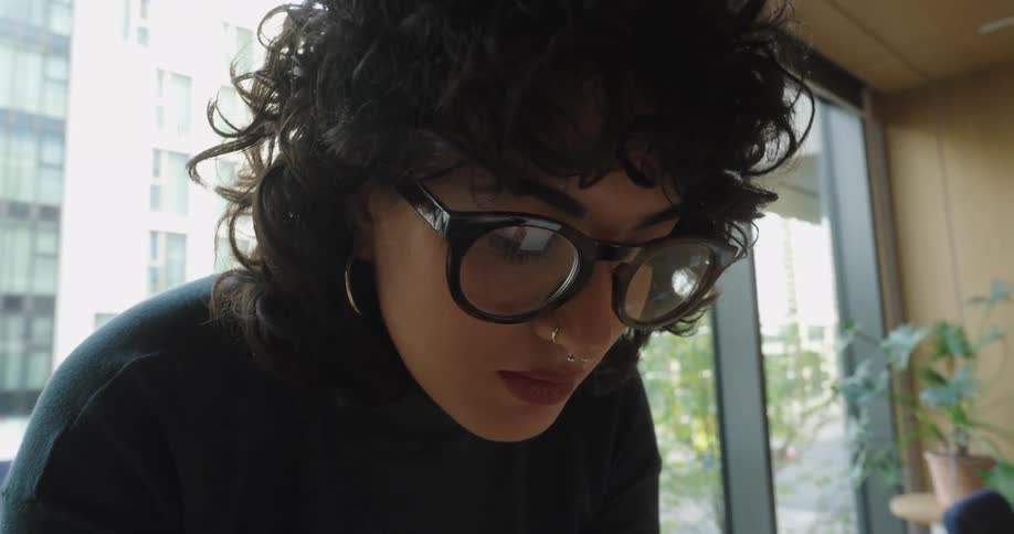 A woman with dark curly hair and large darkframed glasses concentrating intently on something below her with the bright blurred exterior of a modern city building visible through the window behind her