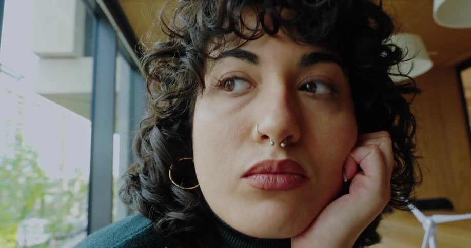 A person with dark curly hair hoop earrings and a septum piercing is shown in a closeup resting their chin on their hand with a thoughtful or slightly worried expression the bright background suggests they are in a modern office or studio setting wit