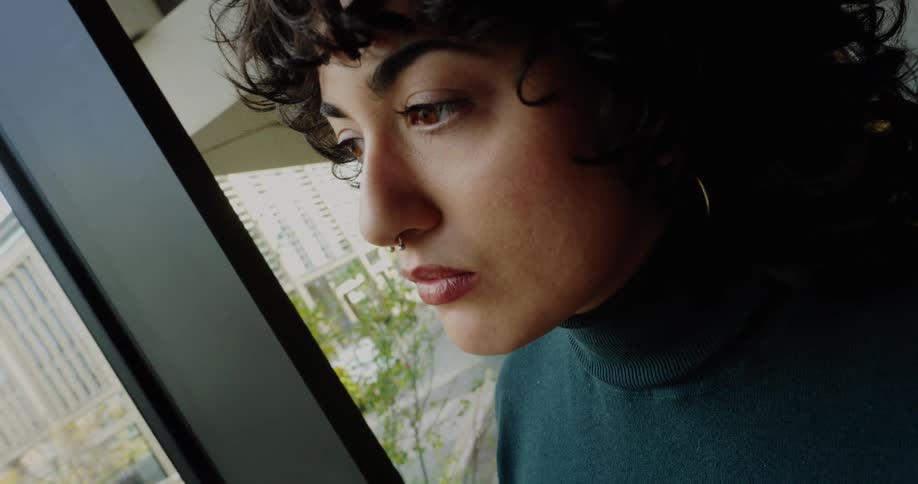 The woman from the previous video is now indoors standing by a large window overlooking a city street she is wearing a dark green turtleneck and lightwash wideleg jeans looking down at her phone or a small object she holds in her hands