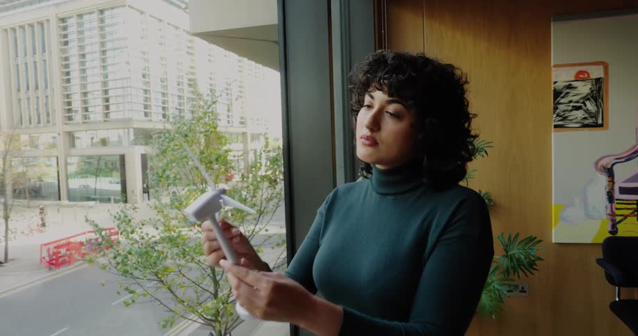 A woman wearing a dark turtleneck and light blue jeans stands by a large office window holding a small white reflex hammershe initially examines the reflex hammer then turns to look out the window at the busy street below closing her eyes briefly