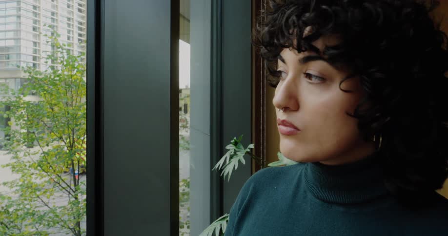 The video focuses on a woman with dark curly hair and a nose ring wearing a dark turtleneck as she stands looking out a large windowthe view through the window shows a city scene with modern highrise apartment buildings and trees with green and yel