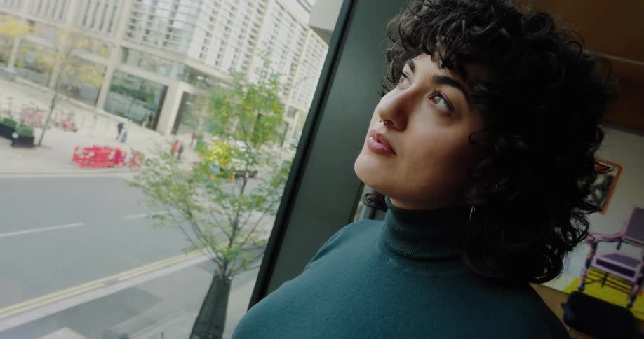 A woman with dark curly hair dressed in a dark turtleneck and jeans stands next to a large window first holding and examining a miniature wind turbine model then slowly looking up and out the window at the cityscape