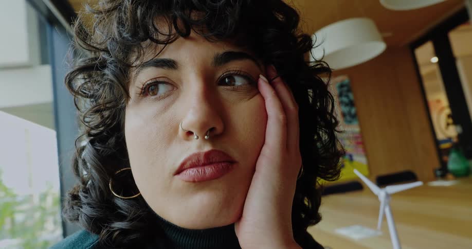 A young woman with curly hair and a nose ring sits behind a conference room desk in an office area appearing deep in thought