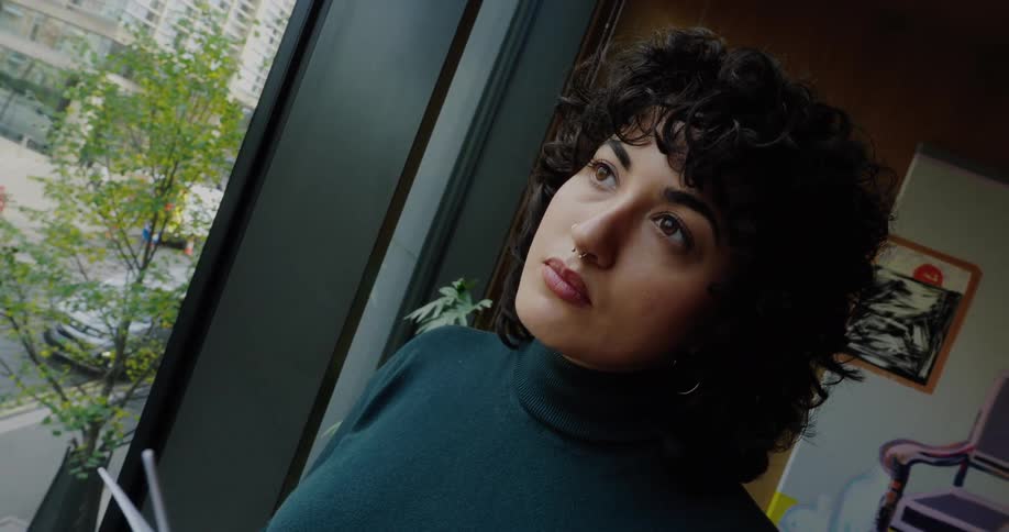 A young black woman with long curly hair stands at midday holding a toy turbine fan while gazing out through a glass window