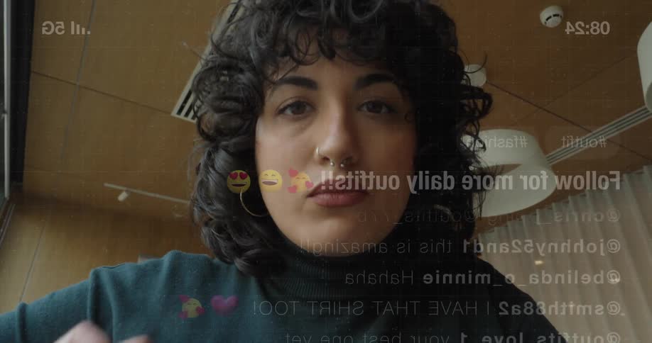A woman with dark curly hair and a nose ring taps a phone screen showing text overlays and obscuring the lens with her hand indoors
