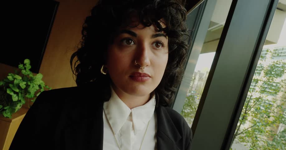 A thoughtful woman with dark curly hair and a nose ring wearing a white shirt and black blazer looks out a large window indoors