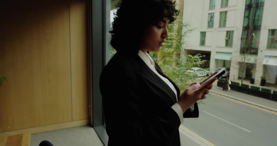 A woman with dark curly hair wearing a white shirt and black blazer stands indoors next to a large window looking down intently at the smartphone held in her hands while the background shows a multistory building across a street