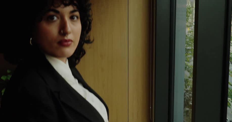 The video starts focused on a carpeted floor before panning up to show a woman standing next to a large window in an office or indoor space she is dressed in a black blazer and white shirtshe looks directly toward the camera with a neutral serious e