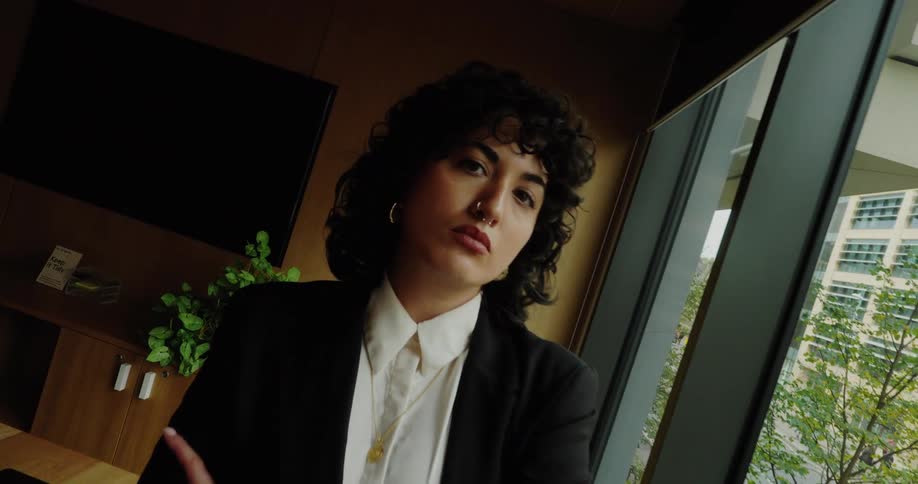 A professional woman in a black suit and white shirt with a nose ring stands with her arms crossed looking out a large window in an office meeting room