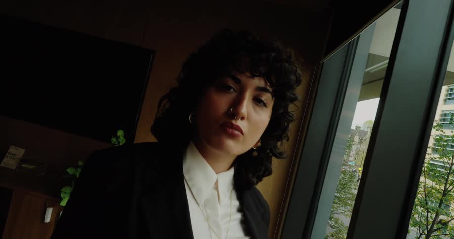 Woman in black blazer looks at cityscape through window in modern room with wooden furnishings