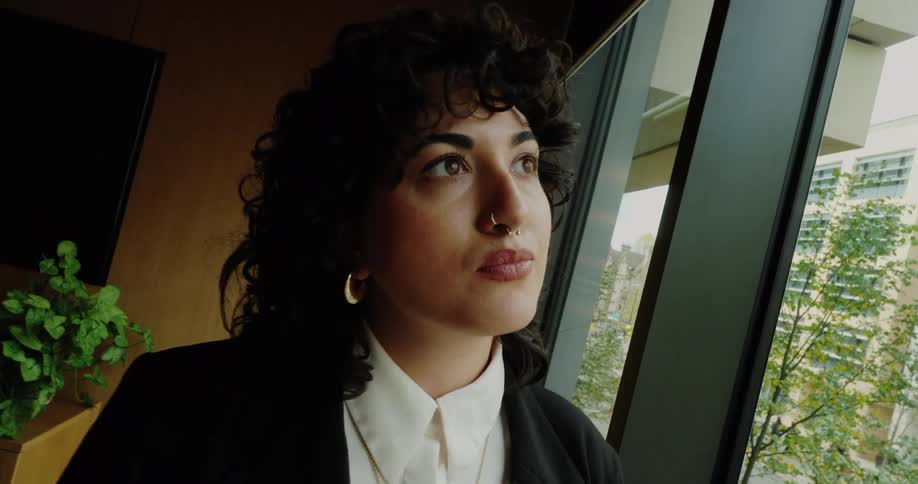 A young woman with dark curly hair wearing a white shirt and black blazer standing by a large window in a modern office as she gazes thoughtfully upward and out toward the city view