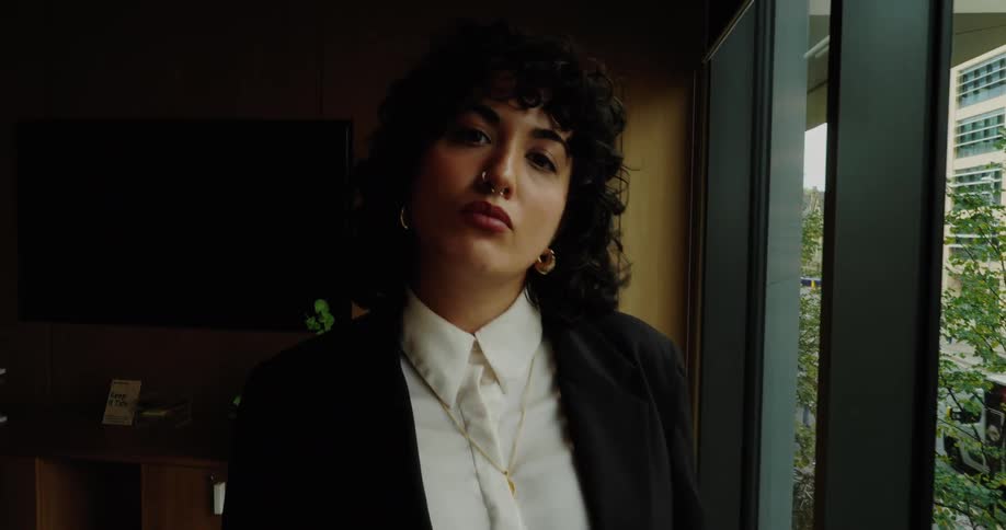 A woman in a black business suit and white shirt stands next to a large interior window overlooking an urban street view and the camera slowly zooms in on her face as she maintains serious eye contact