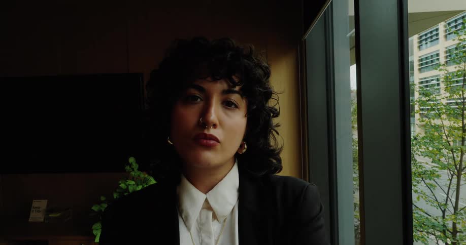 A resolute woman with professional black attire is looking forward with crossed arms inside an office next to a window