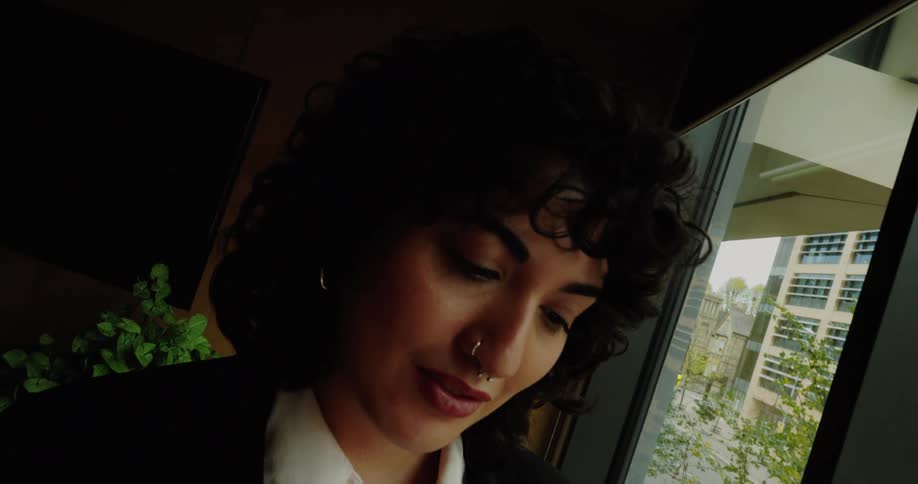 A young person with dark curly hair and a nose ring dressed in a black suit and white shirt stands by a large window using a smartphone they glance up briefly before returning their focus to the device with a multistory building and trees visible out
