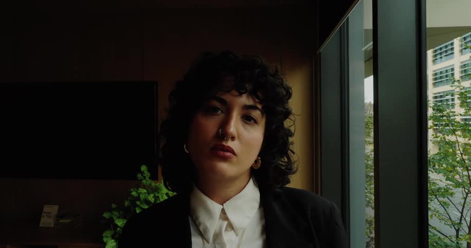 The video begins with a full shot of a woman in a black suit and white shirt standing by a large window in an office settingthe camera then zooms in on her face capturing her direct serious expression