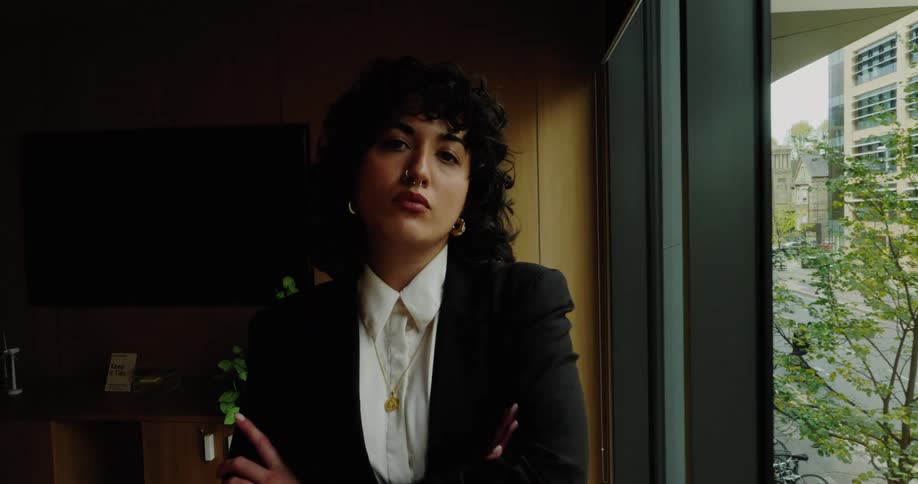 A woman with dark curly hair and wearing a black suit and white shirt stands next to a large window overlooking a city streetshe stares intently into the camera crossing her arms in a professional confident and serious manner