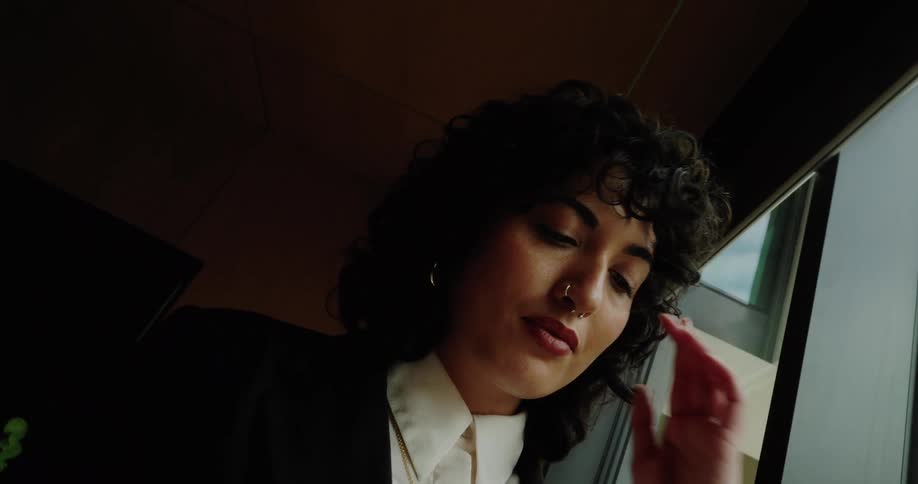 A woman with dark curly hair and a nose ring in a black blazer intently uses her smartphone by a large window indoors