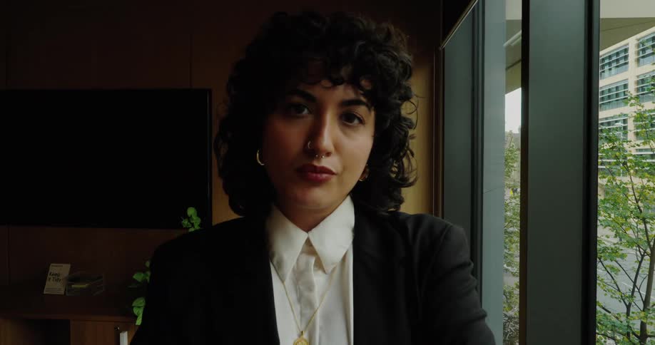 A woman with curly hair and a nose ring stands facing the camera in a professional setting wearing a black blazer over a white collared shirt with her arms crossed while a large window to her right reveals an outdoor area and modern building exterior