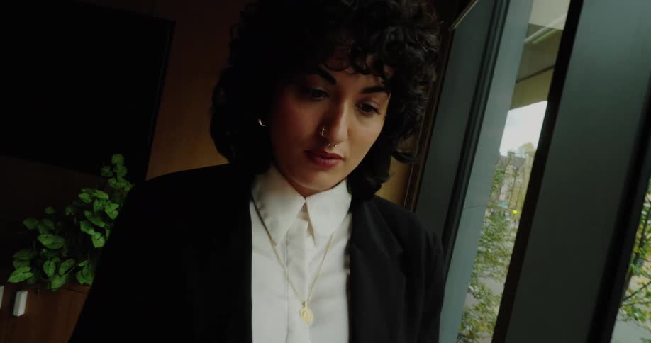 A woman with curly hair and a nose ring in a black suit and white shirt uses her smartphone while standing by a large window indoors