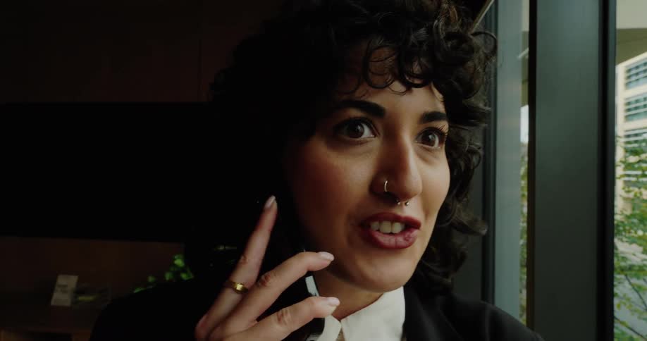 This video clip features a person with dark curly hair and a septum piercing wearing a black blazer over a white collared shirtthey are holding a phone to their ear near a large window appearing to be engaged in a conversation while looking outside a