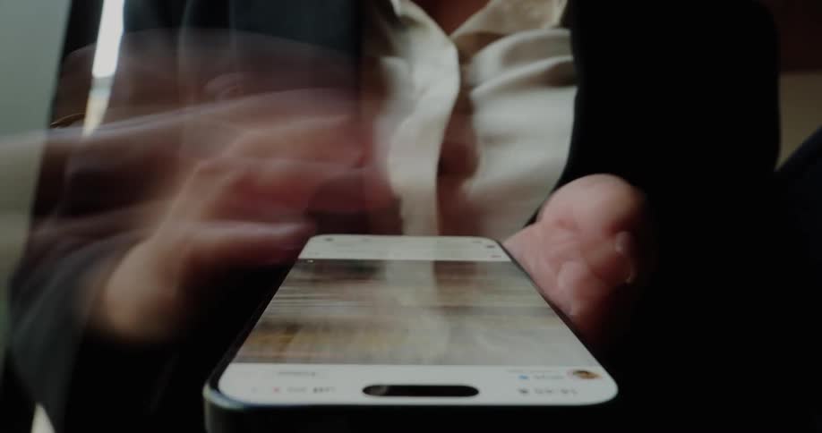 A female is wearing business attire and is looking forward while actively scrolling on a smartphone