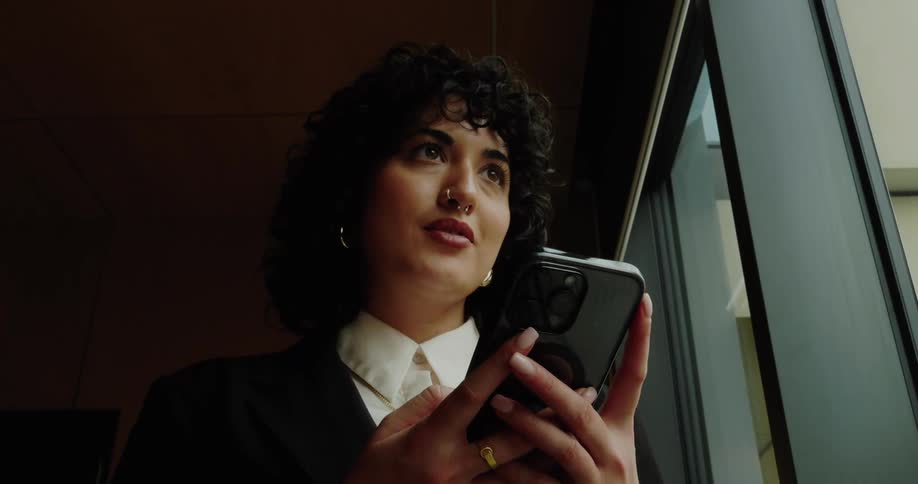 A young black woman stands beside a glass window smiling as she types on her mobile device wearing a nose ring