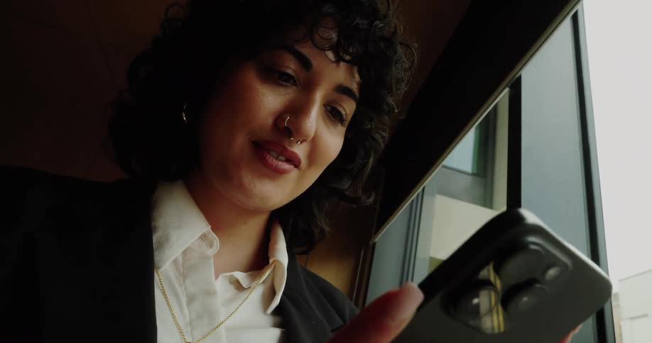 A young black woman stands beside a glass window smiling as she types on her mobile device wearing a nose ring