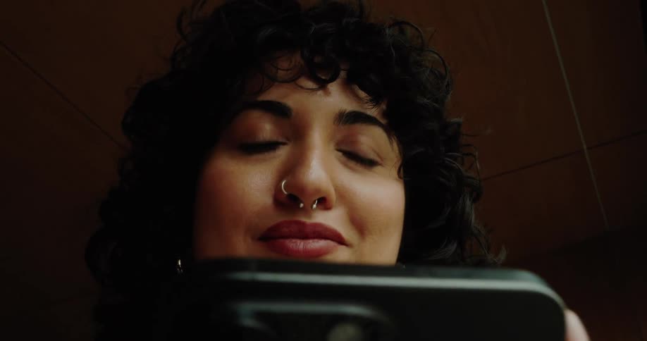 A closeup lowangle shot features a woman with dark curly hair and a septum piercing looking down at a smartphone she is holding she closes her eyes and then looks up suggesting a moment of thought or reaction to what she is viewing on the screen