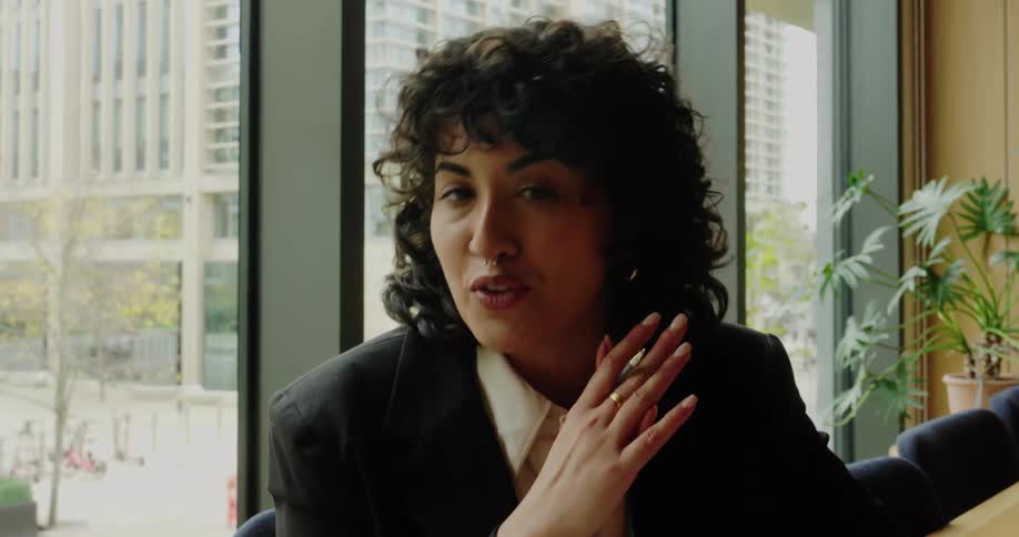 The woman seen in the first video is now sitting at a table indoors possibly in an office or a cafe with large windows she is wearing a black blazer and white shirt and appears to be speaking animatedly gesturing with both hands while holding a pen