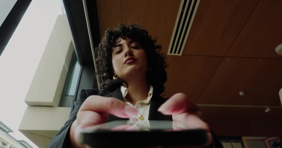 Young adult woman texting on a smartphone indoors next to a window during day time