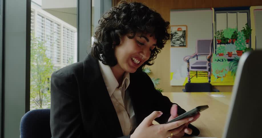 The video captures a person with short dark curly hair wearing a black blazer over a light shirt seated at a desk and smiling while looking at their phone they have a nose ring and appear relaxed and happyin the background there is a bright windo