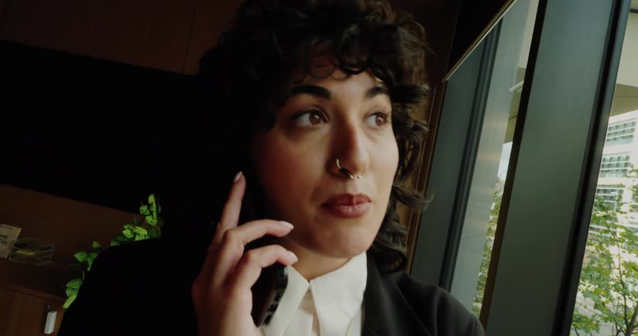 A stylish woman with dark curly hair and a nose ring wearing a black suit and white shirt is standing by a large window talking on a cell phoneshe is indoors in an office or corporate setting with the camera zooming in as she smiles and continues h