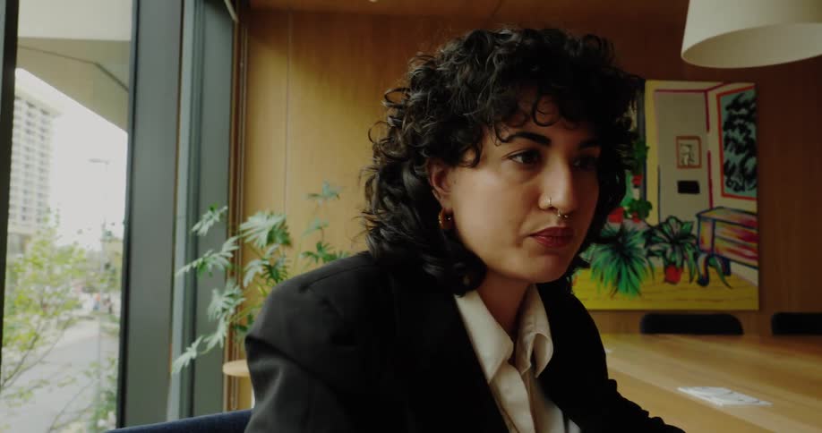 The video begins with a closeup of a persons hands with light nail polish and a ring typing quickly on a laptop keyboard at a long wooden tablethe camera then pans up to show the person who has curly dark hair and a nose ring wearing a black blaz