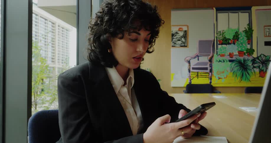 A woman sits by a large window texting on her smartphone in a bright modern office