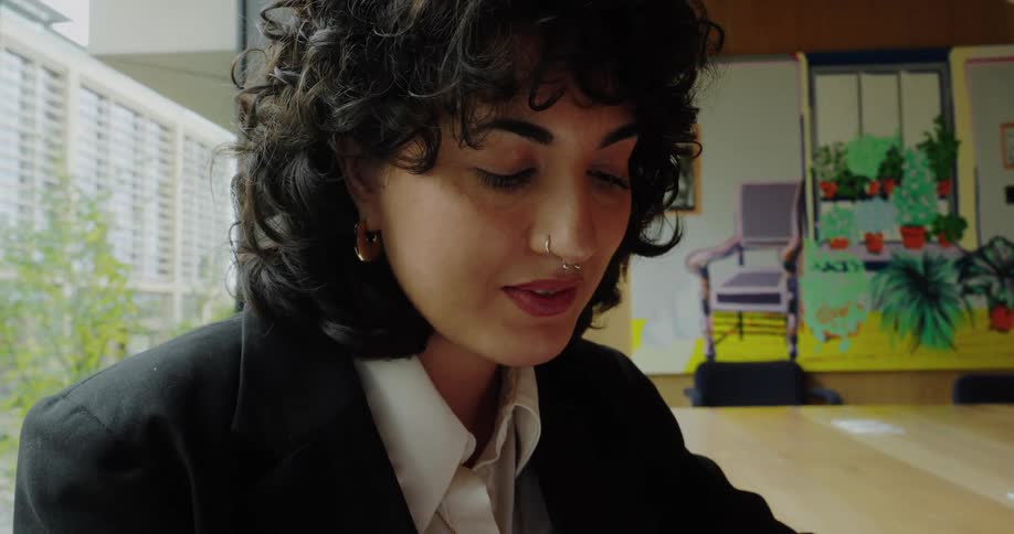 A woman dressed in a business suit holds a smartphone with a neutral expression looking forward while sitting at a wooden desk with a notebook and pen
