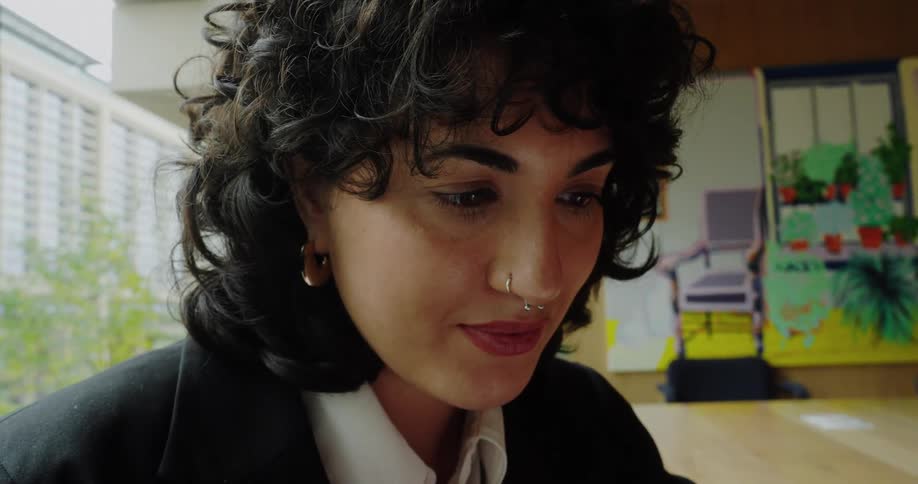 A womans hands using a smartphone while she wears a black blazer and white shirt and then quickly shifts to a closeup of her face as she intently focuses on the device with a slight smile in an indoor setting featuring abstract artwork in the back