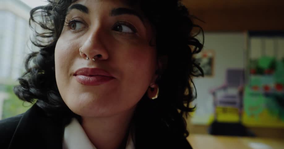 The video is a closeup on the face of a person with dark curly hair full eyebrows and light makeup they have a double nose piercing a septum ring and a stud and small hoop earringsthey initially look down and then briefly glance up and to the s