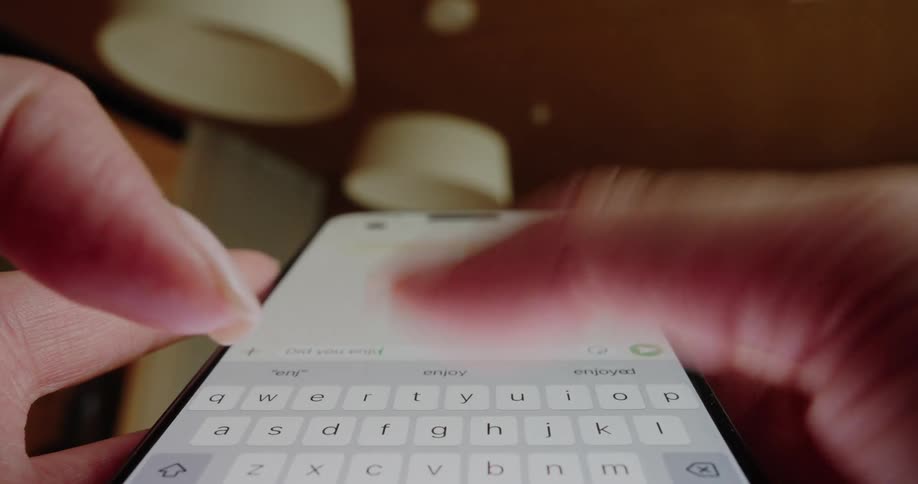An extreme closeup lowangle shot focuses directly on a persons fingers rapidly typing a message on the onscreen keyboard of a smartphone with two outoffocus white light fixtures visible in the background