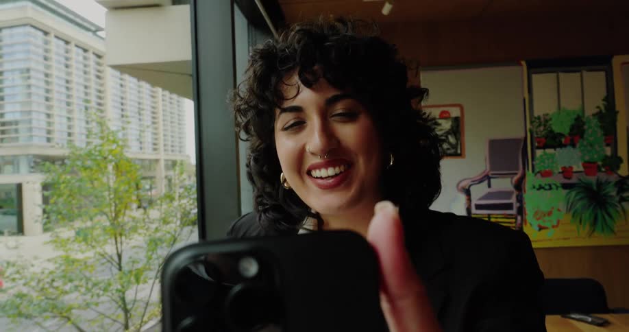 A young woman wearing a jacket and a nose ring stands beside a window smiling as she excitedly makes a video call on her mobile device