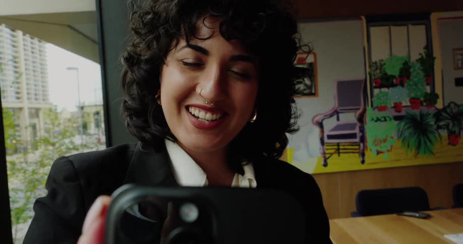 A woman from the arms up is seen waving and talking to a smart phone appearing to be on a video chat in a room
