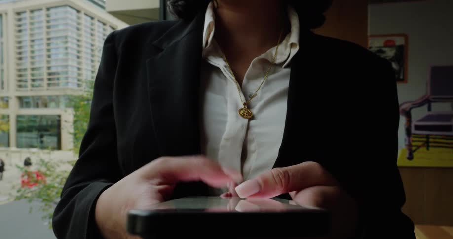 A professional woman in a blazer types on her smartphone in front of a window