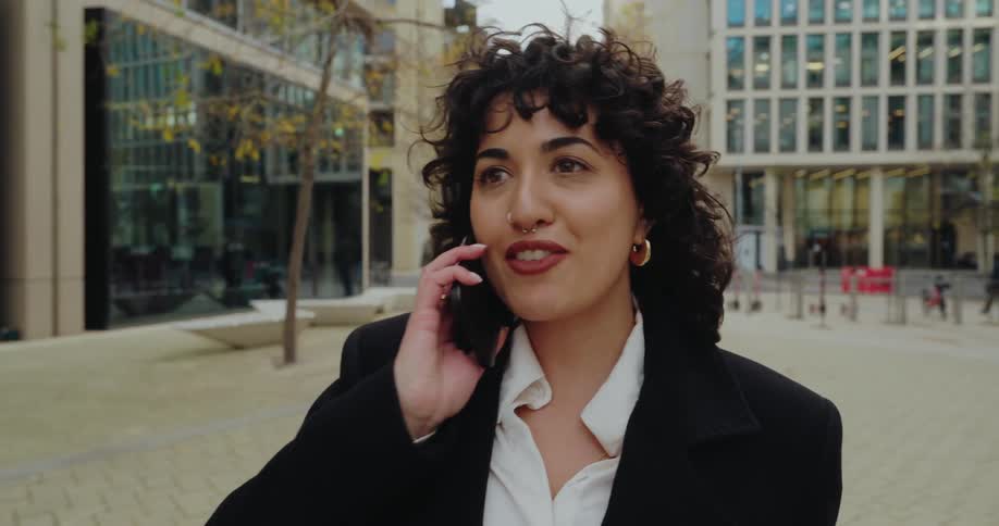 A young woman with dark curly hair and a nose ring is standing outside a modern building complex she is wearing a black blazer over a white shirt she is holding a smartphone up to her ear and appears to be in the middle of a phone call