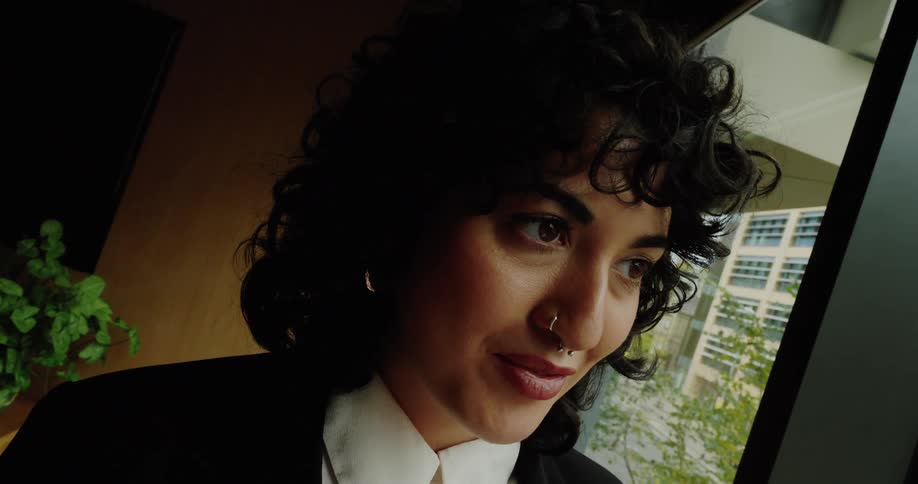 A woman with dark curly hair and a nose ring dressed in business attire stands next to a large window in a conference room looking down at her mobile phone before looking up and smiling warmly