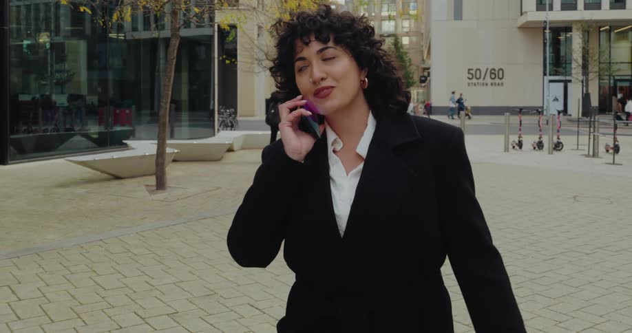 A professional woman with curly hair wearing a black coat and white shirt walks along a paved modern urban plaza while talking on her phonein the background there is a building signed 5060 station road and several electric scooters are lined up