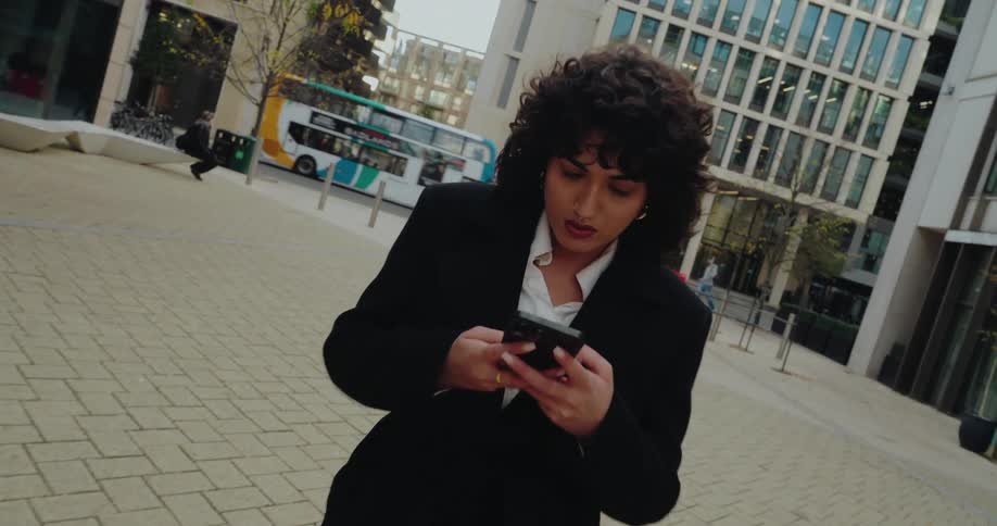 A woman with dark curly hair dressed in a black overcoat and a white shirt on a modern paved urban plaza looking down and interacting with a smartphone in her hands while a doubledecker bus passes in the background
